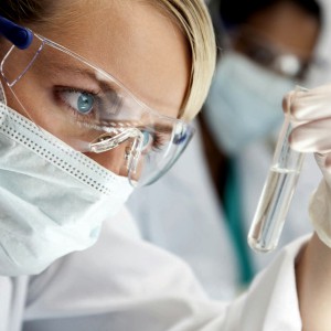 A blond medical or scientific researcher or doctor using looking at a clear solution in a laboratory with her Asian female colleague out of focus behind her.